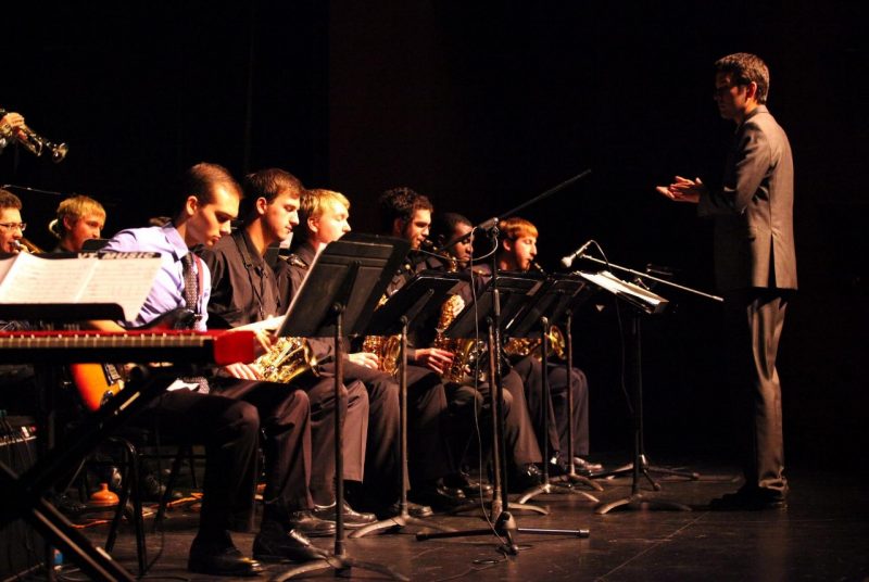 Performers in the jazz ensemble sit on stage and play, with their director in front of them.