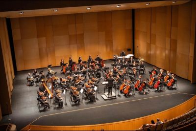 Members of the Virginia Tech Philharmonic Orchestra sit on the stage of the Moss Arts Center with their instruments.