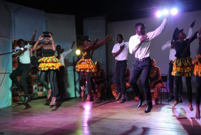 Young people dressed in black pants/white shirts and black top/leggings and colorful skirts dance on stage.