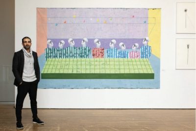 A man stands in a gallery beside a large, colorful artwork. The artwork features a row of stylized human figures with identical faces and striped clothing, seated behind a green grid-like surface against pastel blocks of purple, pink, blue, and yellow. Small multicolored circles float above them. To the right, two smaller framed pieces display minimalist objects resembling metal tools or pins mounted on white backgrounds. The man wears dark pants, a light gray sweater, and a dark jacket, with one hand in his pocket, looking toward the camera.