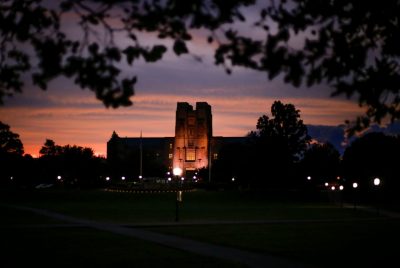 A large stone building with a tall central tower is illuminated at dusk, framed by dark tree branches in the foreground. The sky glows with deep orange and purple hues as scattered lights line a pathway across a wide lawn leading toward the building.