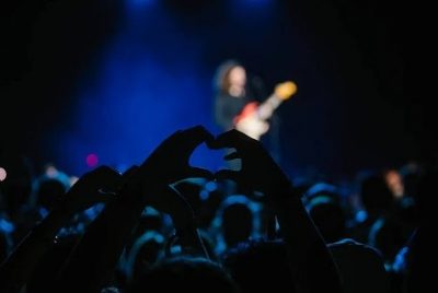 Audience members form a heart shape with their hands during a concert, with a musician playing guitar on a brightly lit stage in the background.