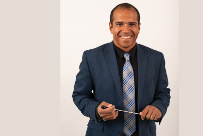 Armand Hall, a man dressed in dark suit and plaid tie, holds a conductor's baton