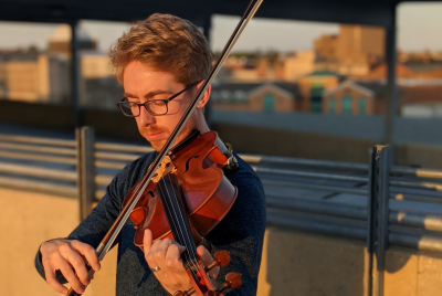 Gavon Peck, a young man, stands outdoors and plays a violin with the reflection of buildings in the background