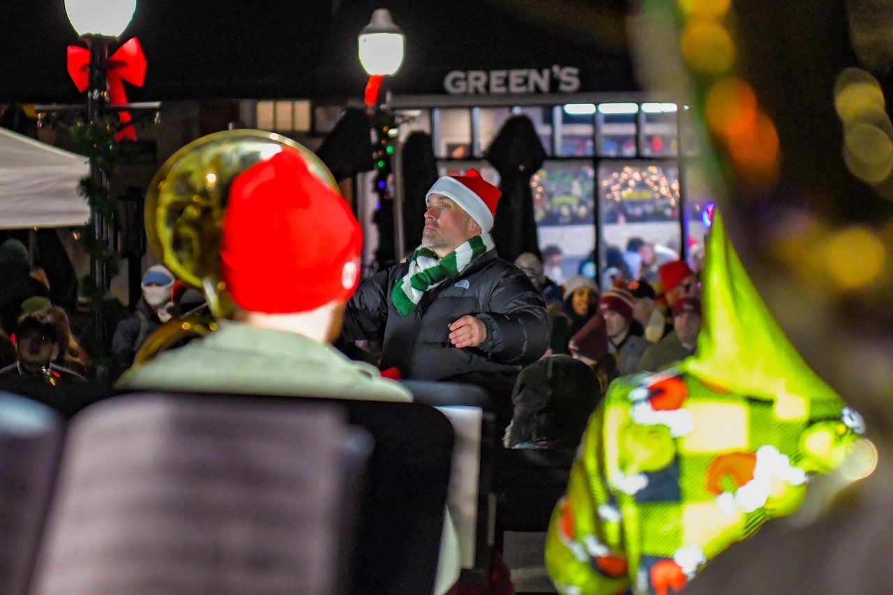 Band director wearing Santa hat and the back of students playing tubas at night.