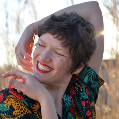 Rachel Rugh, short brown hair, wearing a colorful red, yellow, teal, and black shirt, smiles and holds her hands around her face