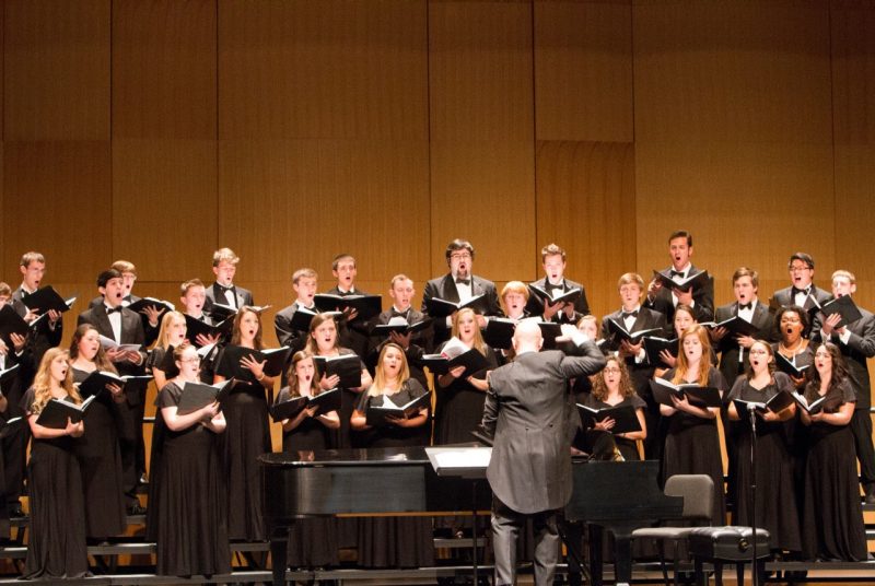 Male and female chorial students,  dressed in black, perform on stage with their conductor.