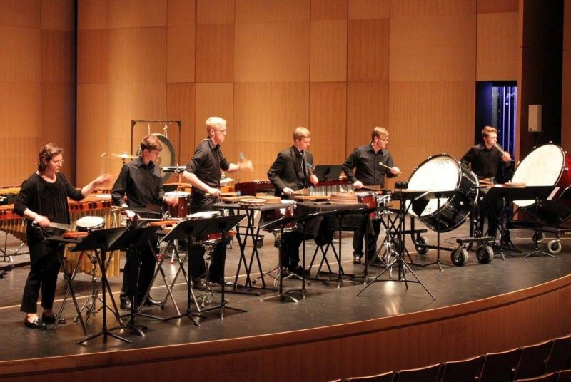 Six musicians dressed in black on stage, playing mallet instruments, bass drums, and other percussion instruments