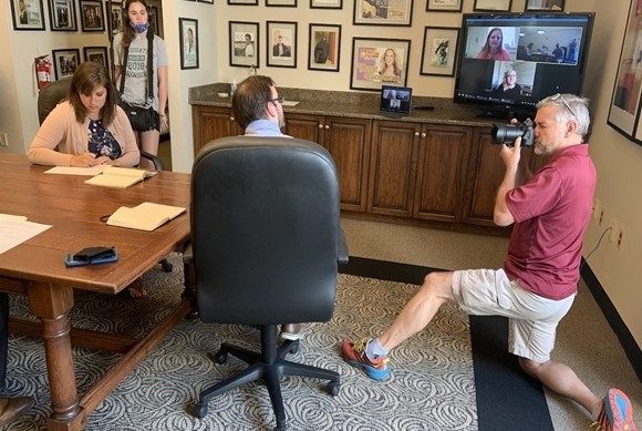 Four people in a room with a table. One man is knelling down to take a photo. The walls are colvered with framed drawings, and there is a computer display with a Zoom meeting on the screen.