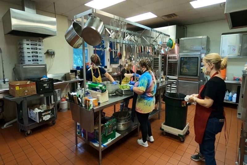 Four people wearing face masks in an industrial kitchen preparing food at a station with pots, pans, and cooking utensils overhead.