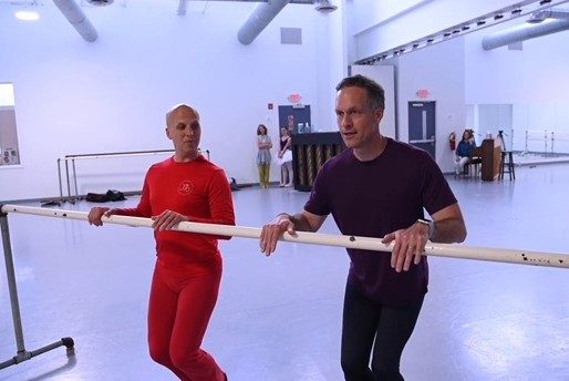 Two men stand at a barre in a ballet studio, with three other people in the background near a piano, watching.