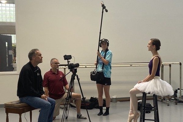 A young women in a white tutu sits on a stool facing a camera. Two men sit behind the camera on a bench, and another person stands nearby holding a boom microphone. There are a set of ballet barres in the background.