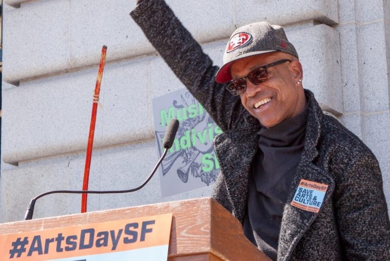 A man wearing sunglasses, cap, and drak shirt and jacket, stands behind a podium with one arm raised. The sign on the podium has the hashtag #ArtsDaySF.