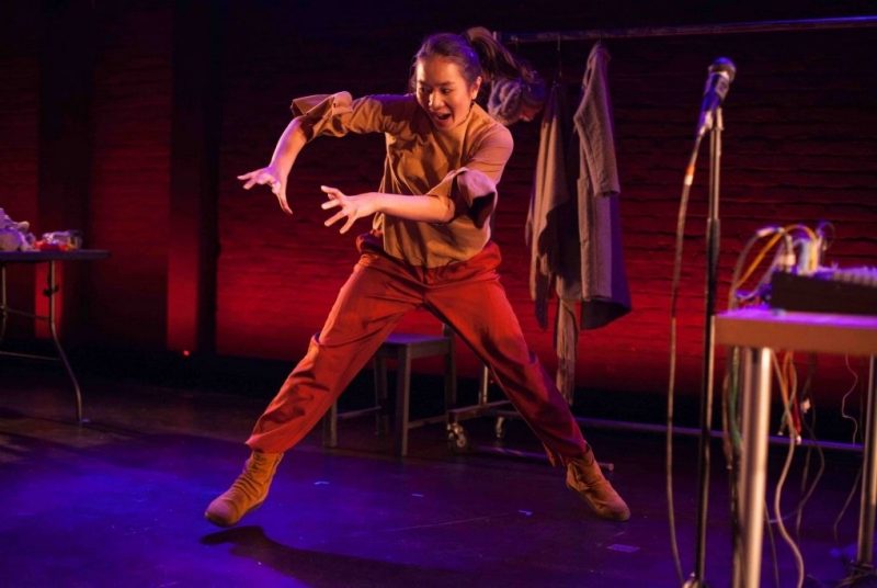 An indigenous woman wearing reddish-brown shirt and pants is in mid-performance, mid-movement. She is on a sparsely-furnished staged lit with dynamic red and blue lights.