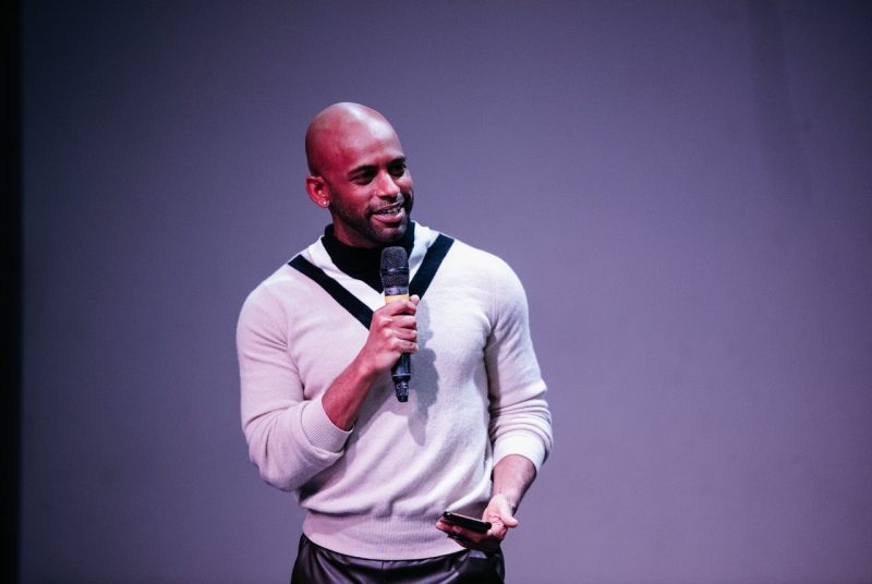 Joseph Hall stands, holding a microphone, wearing a white pullover sweater.