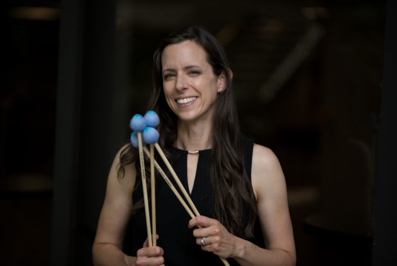 A woman in a sleeveless dark top stands holding two percussion mallets in each hand.