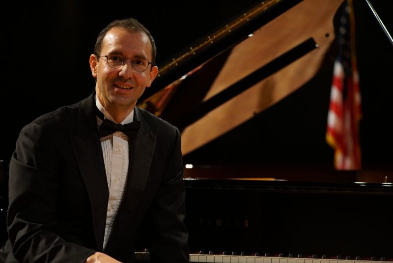 A man with glasses, wearing a tuxedo with a bow tie, sits in front of a grand piano