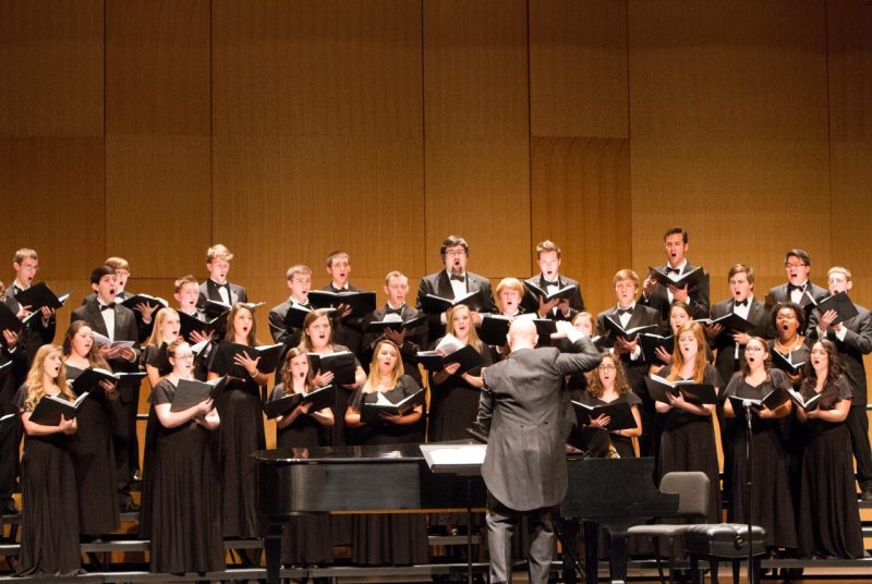 College choir members, male and female, dressed i black, perform on stage, standing behind a grand piano and the conductor