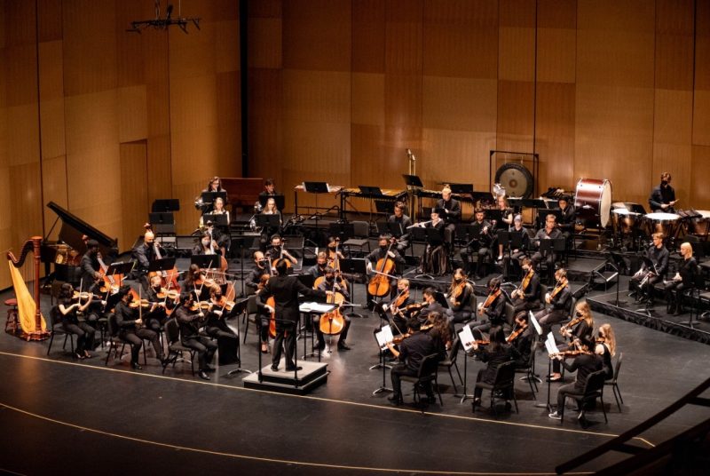 Virginia Tech students in the VT Philharmonic orchestra are on stage at the Moss Arts Center, dressed in black, performing with the conductor, also dressed in black standing on the podium.
