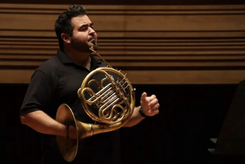 Chris Castellanos, dark hair, wearing a dark shirt, holds a French horn with one hand inside the bell of the instrument.