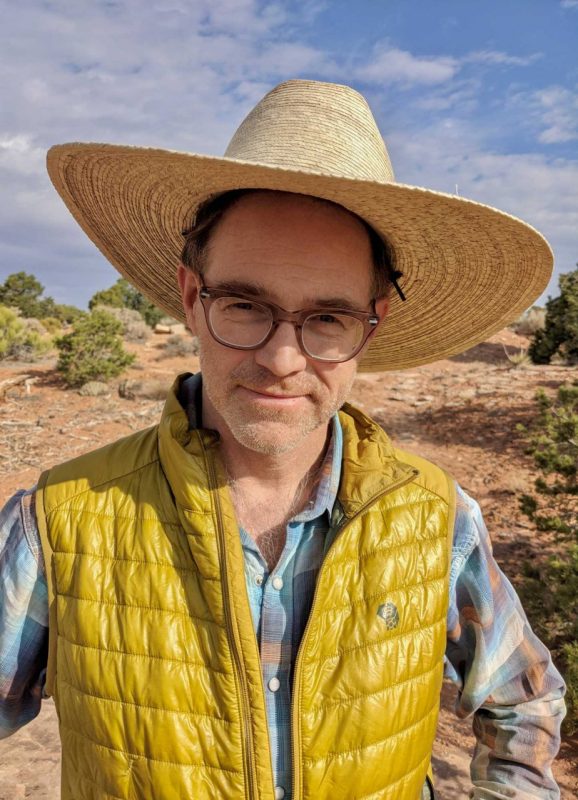 A man wearing glasses, a larte straw hat, a plaid shirt, and yellow vest, stands in the desert.