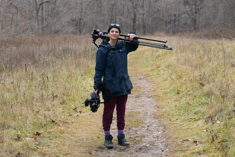 A woman stands in a field holding a camera tripod behind her neck, and a movie camera in one hand.