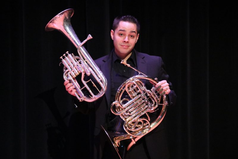 A man in a dark shirt and jacket holds a French horn in one hand and an alto horn in the other hand.