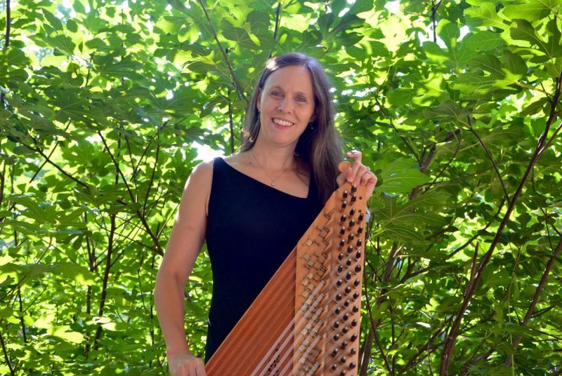 Anne Elise Thomas, wearing a black dress, stands outside in front of some trees, hold a qanun,  a 78-stringed zither. instrument.