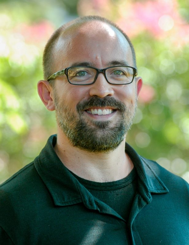 A man wearing dark rimmed glasses and a dark green shirt smiles