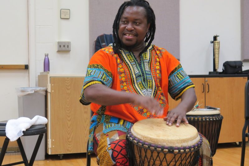 Oto Koju wears a colorful orange and blue shirt and plays a West African drum