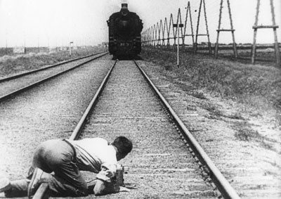 A black and white still from a movie showing a man crawling over a railroad track while a locomotive heads down the track.