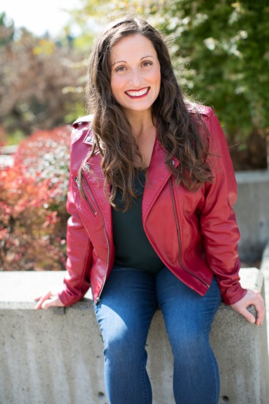 A smiling woman in a red jacket and jeans, its outside on a concrete wall.