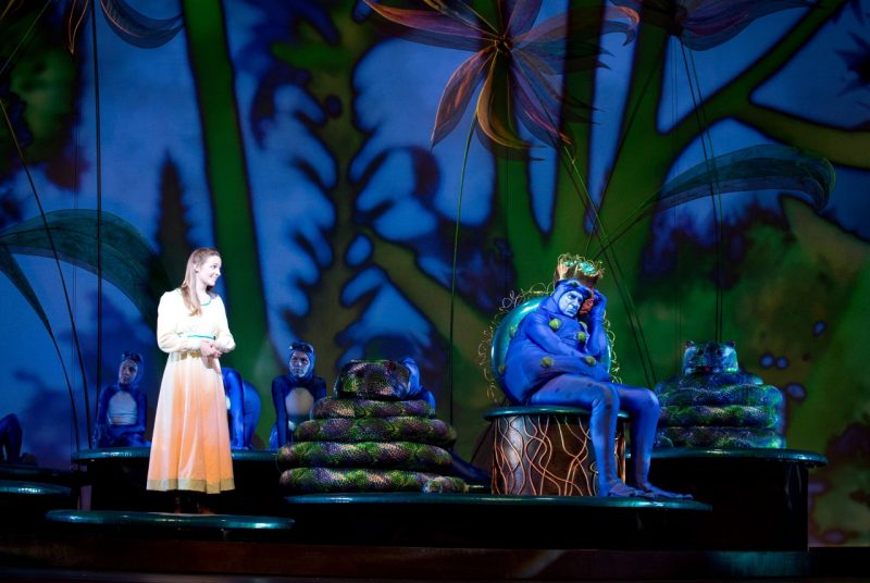 A blue frog prince character sits while a young woman looks on from the production of "The Three Feathers" children's opera