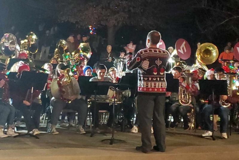 Musicians dressed in Christmas and holiday attire, stand outside and play low brass instruments.