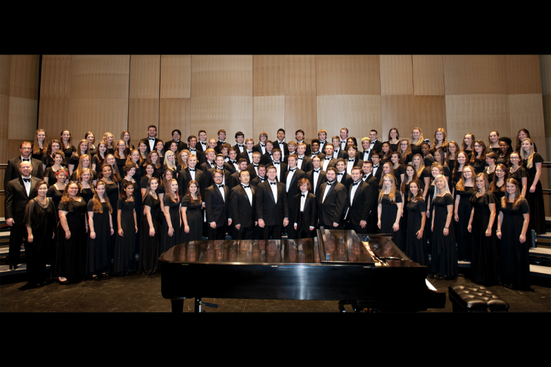 Members of the Virginia Tech choirs dressed in black, stand on stage behind a grand piano