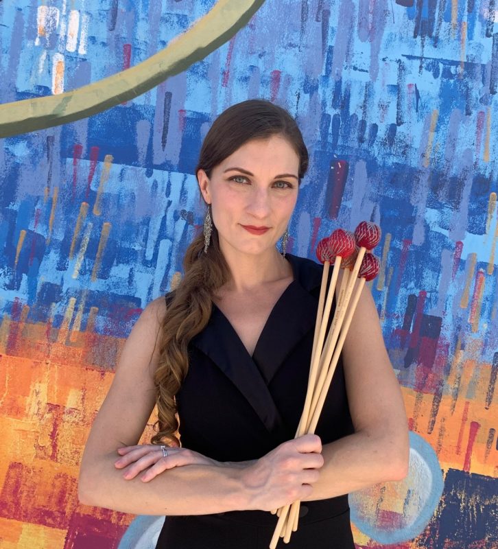 Karli Vina, with long brown hair, wearing a black dress, stands in front of a colorful blue and orange background holding percussion mallets.