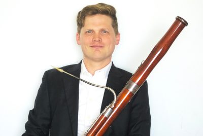 A man with short, light brown hair, dressed in a white shirt and black jacket, holding a bassoon.